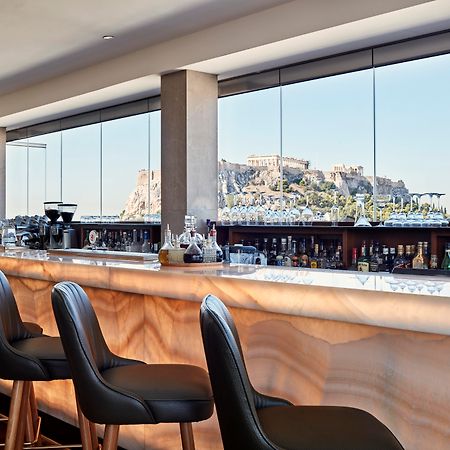 فندق إلكترا متروبوليس أثينا المظهر الخارجي الصورة The photo features a modern bar area with a sleek design. It showcases a long bar counter made of a light-colored material, possibly a type of stone, that has a smooth, polished finish. Behind the bar, there are shelves lined with various bottles, su