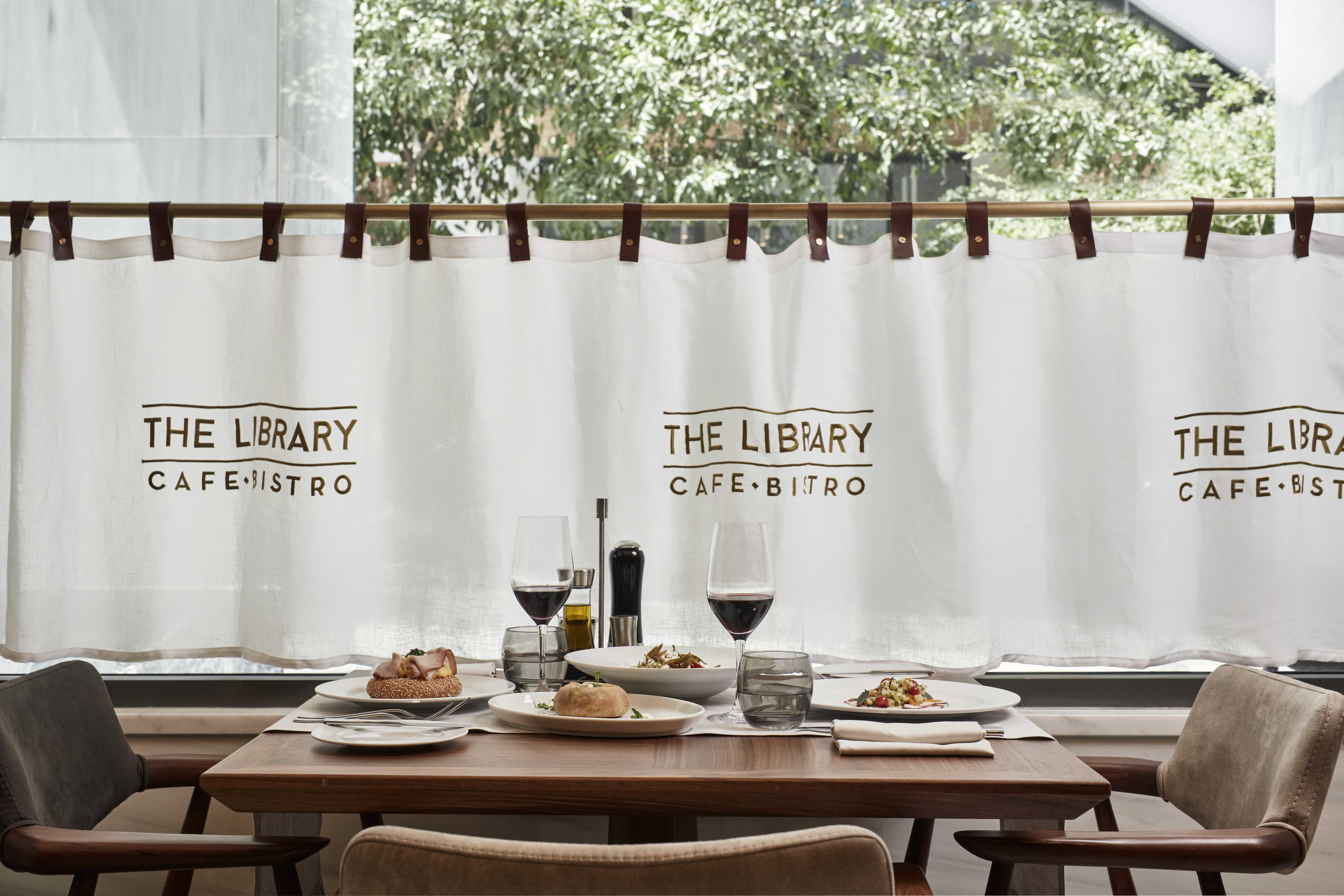 فندق إلكترا متروبوليس أثينا المظهر الخارجي الصورة The photo shows a cozy dining setting in a café or bistro named "The Library Café-Bistro." There is a table with several plates of food, including what appears to be a salad and some baked items. Two glasses of red wine are prominently placed on the 