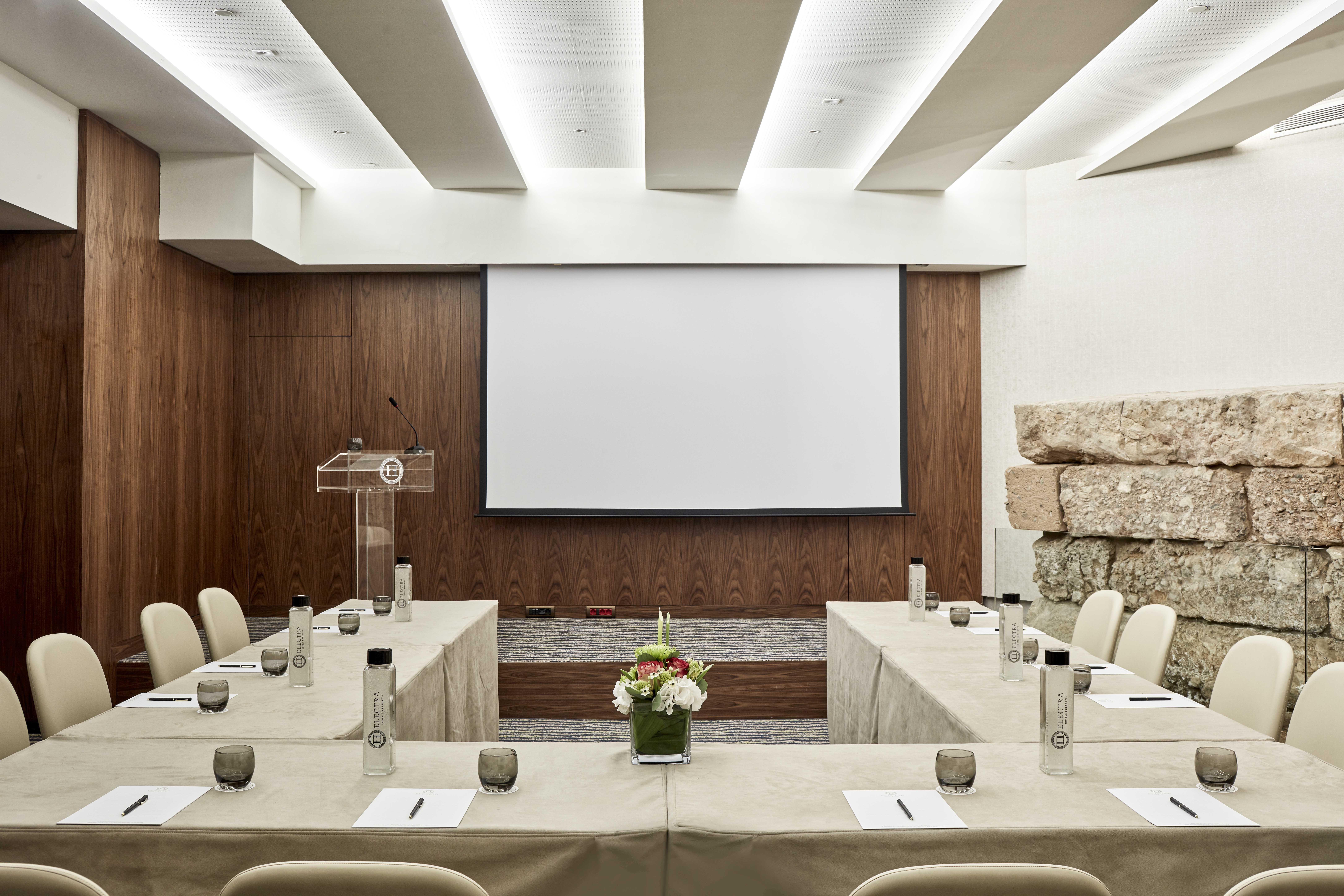فندق إلكترا متروبوليس أثينا المظهر الخارجي الصورة The image shows a modern conference room setup. The room features a large screen at the front, likely for presentations. There are several tables arranged in a U-shape, covered with light-colored tablecloths, and each table has glasses and notepads. 