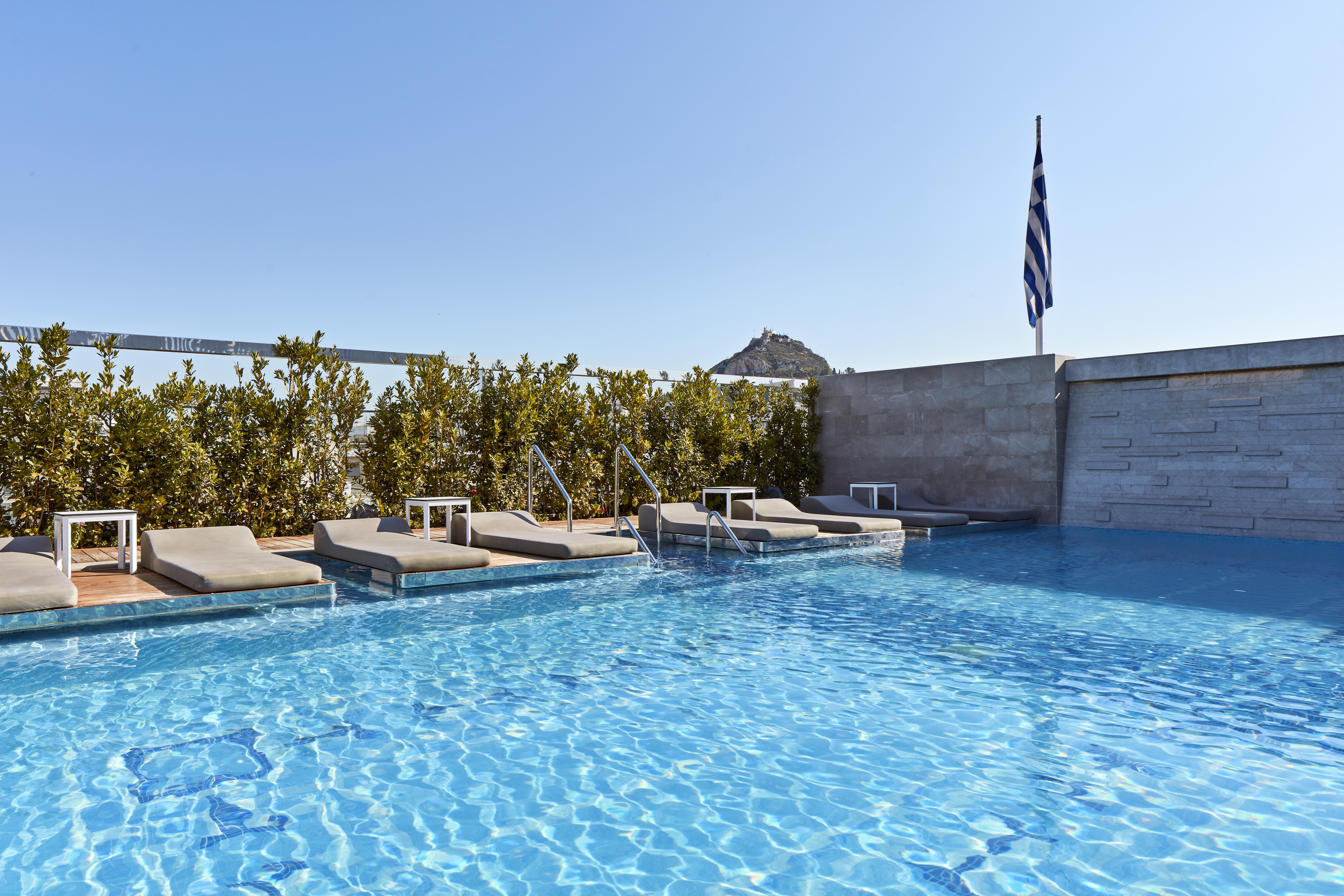 فندق إلكترا متروبوليس أثينا المظهر الخارجي الصورة The photo shows a rooftop swimming pool area. The water in the pool is clear and inviting, reflecting the blue sky above. There are several lounge chairs positioned around the pool, suggesting a relaxing atmosphere. In the background, there are green