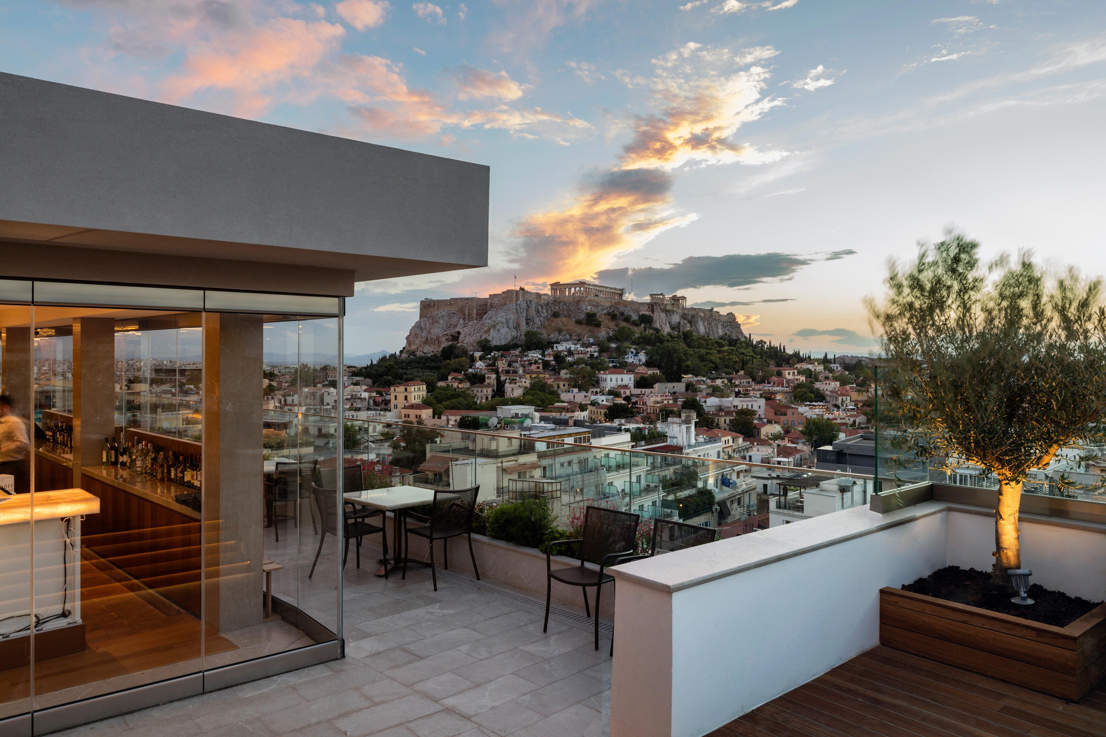 فندق إلكترا متروبوليس أثينا المظهر الخارجي الصورة The photo shows a rooftop terrace with modern furnishings, including tables and chairs, set against a stunning backdrop. In the distance, there is a prominent hill with historic architecture, likely an ancient monument, suggesting it may be an iconic