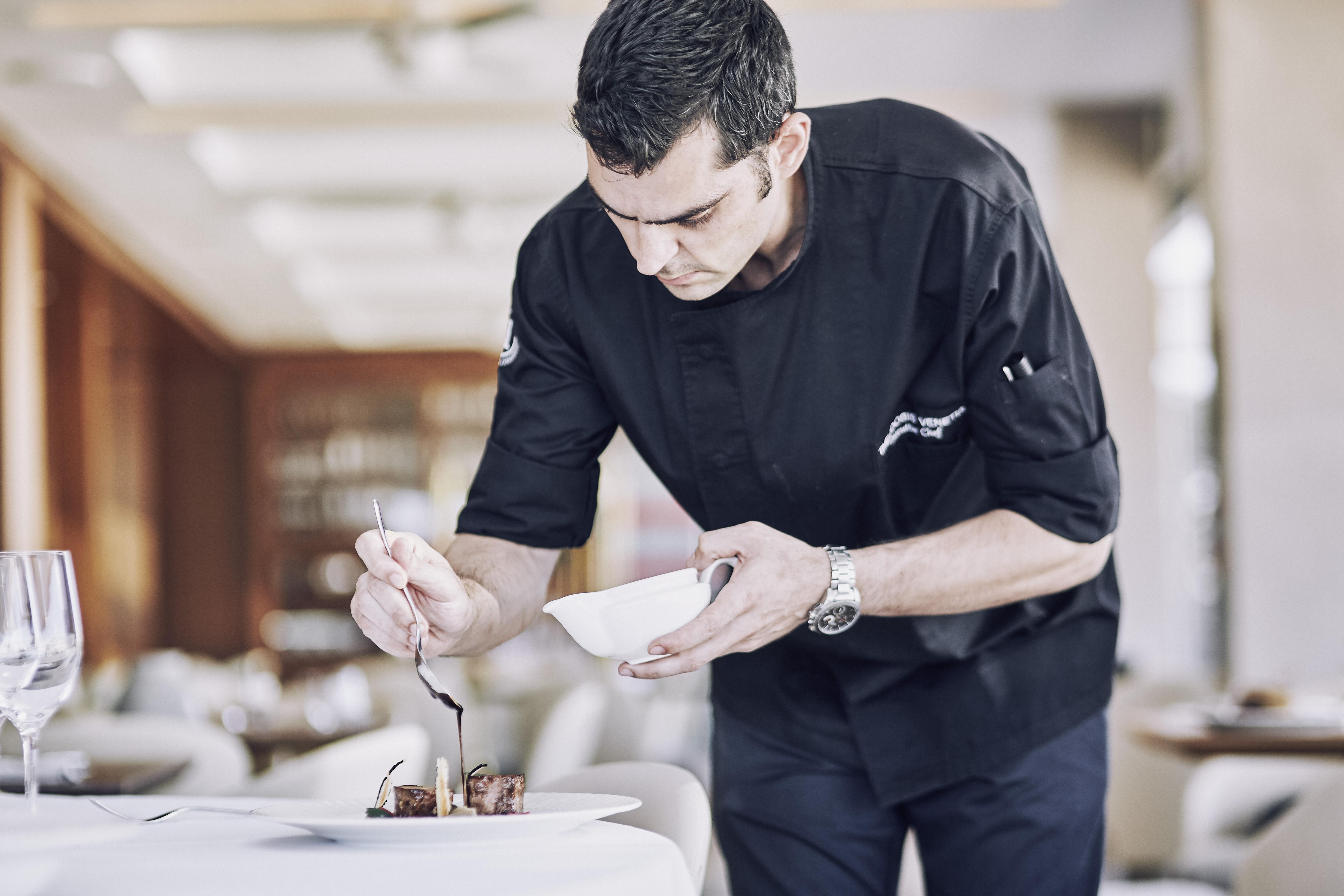 فندق إلكترا متروبوليس أثينا المظهر الخارجي الصورة The photo shows a chef in a black chef's jacket carefully plating a dish. He is leaning over a table, focused on adding a finishing touch to the presentation with a spoon. The dining area has a modern and elegant ambiance, with a soft focus on the ba