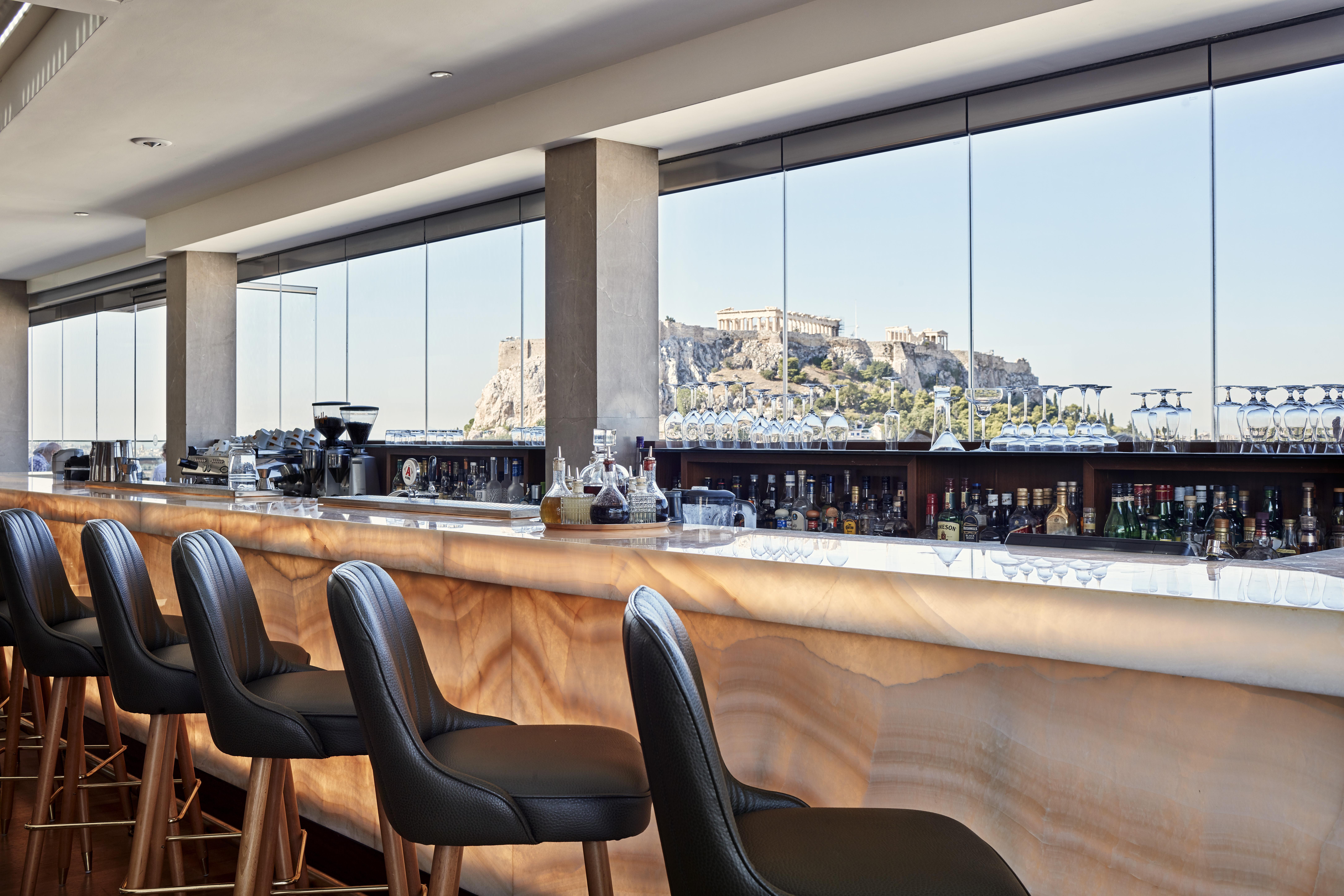 فندق إلكترا متروبوليس أثينا المظهر الخارجي الصورة The photo features a modern bar area with a sleek design. It showcases a long bar counter made of a light-colored material, possibly a type of stone, that has a smooth, polished finish. Behind the bar, there are shelves lined with various bottles, su