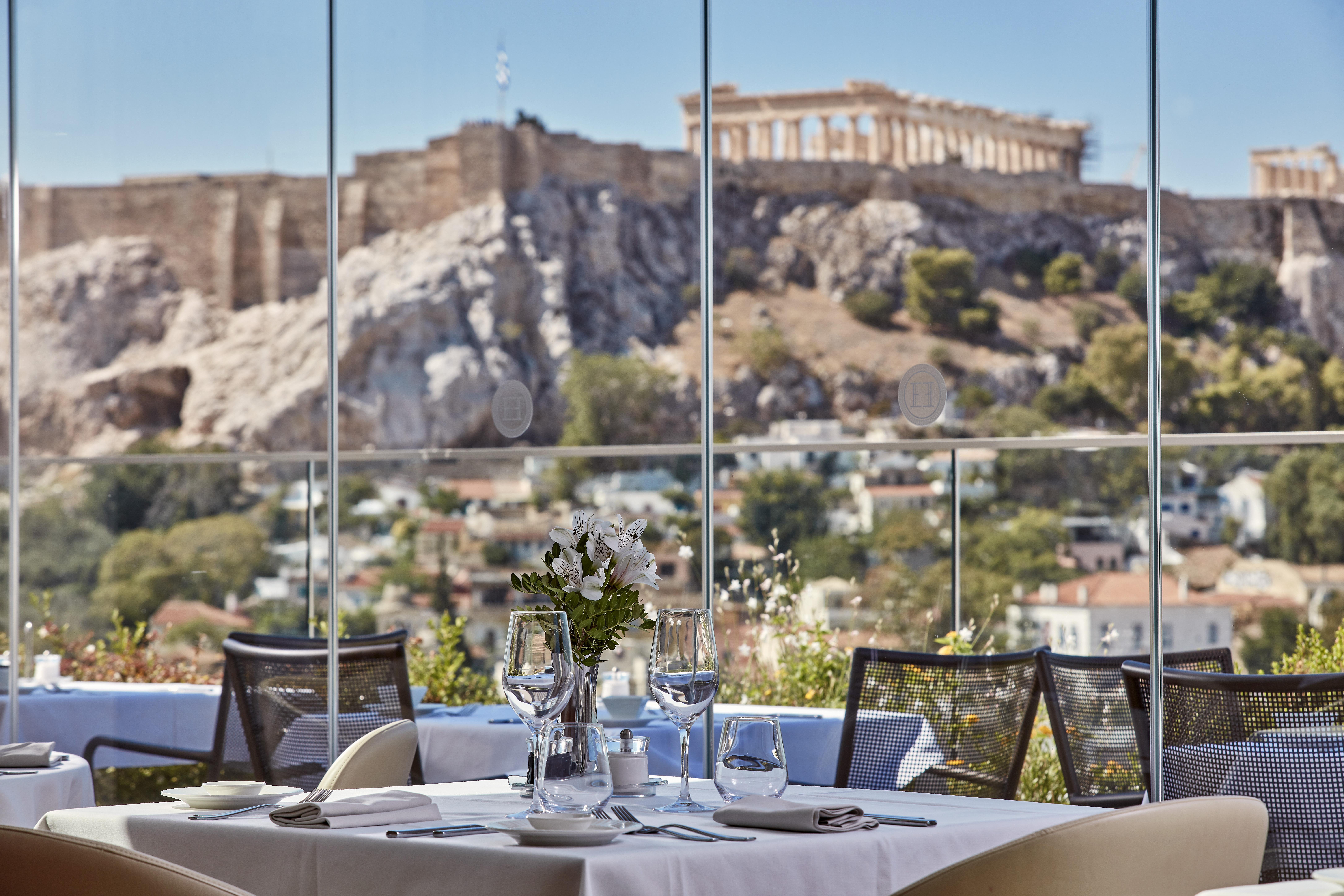 فندق إلكترا متروبوليس أثينا المظهر الخارجي الصورة The photo shows a beautifully set dining table with white tablecloths and elegant glassware. In the background, there's a stunning view of a historic site, likely the Acropolis in Athens, Greece. The scene captures both the modern dining experience a