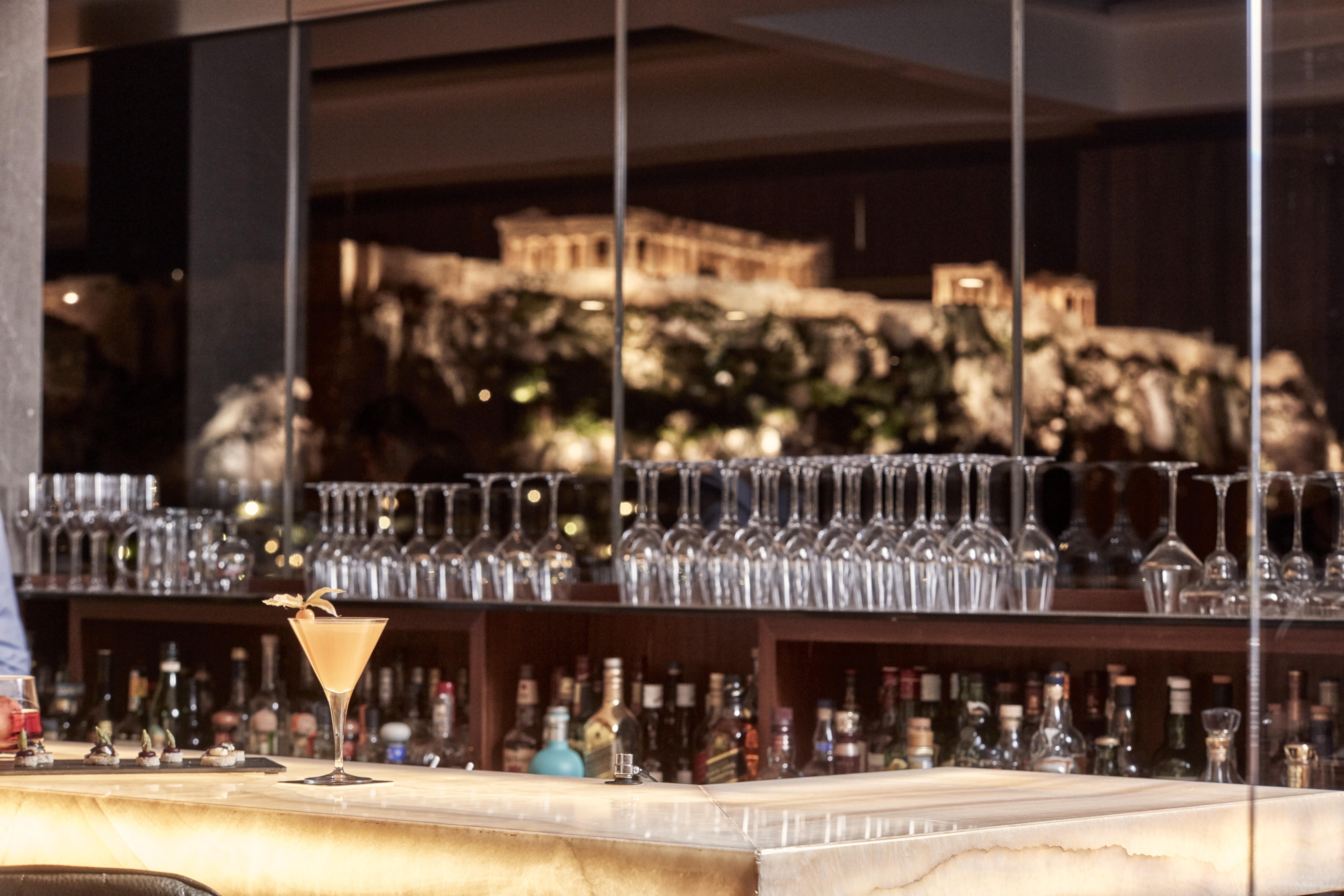 فندق إلكترا متروبوليس أثينا المظهر الخارجي الصورة The photo showcases a sophisticated bar setup, featuring a marble counter with a cocktail placed elegantly on it. Behind the bar, there are numerous glassware items neatly arranged on shelves, alongside various bottles of alcohol. In the background, 