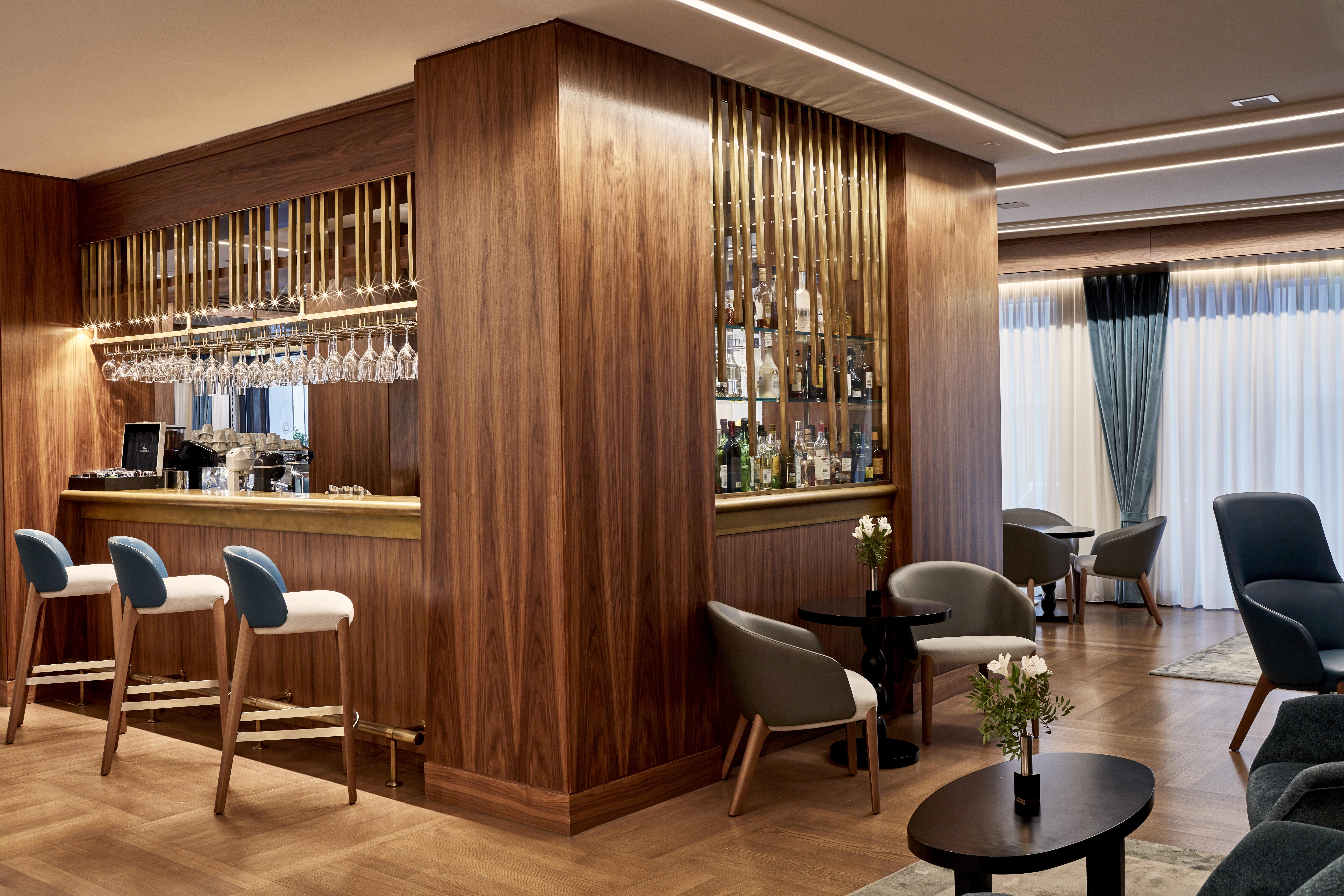 فندق إلكترا متروبوليس أثينا المظهر الخارجي الصورة The photo depicts an elegantly designed bar area. The bar itself features a wooden structure with a display of various bottles and glassware, highlighted by warm lighting. In front of the bar, there are tall white and gray stools. The flooring is woo