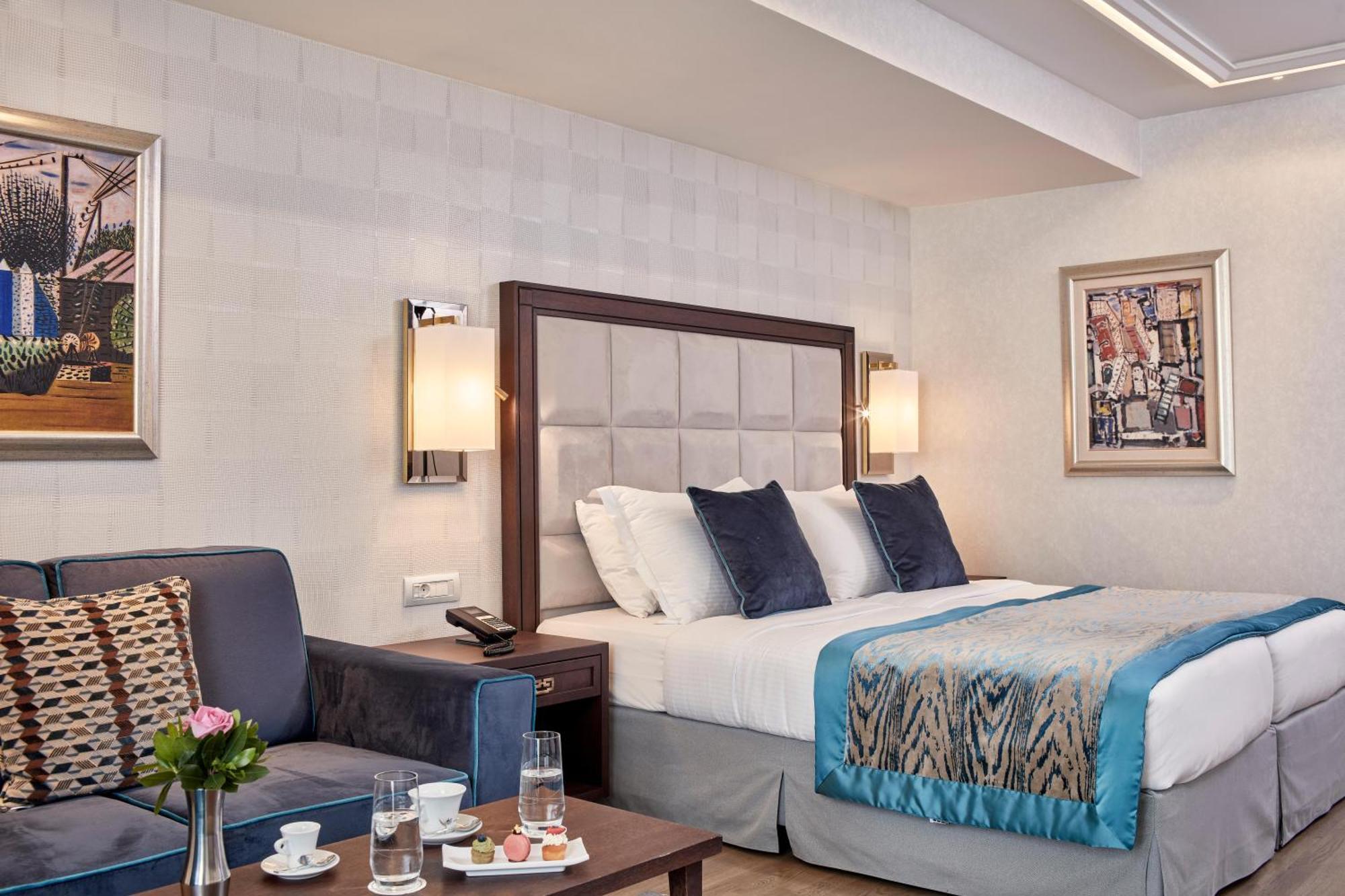 فندق إلكترا متروبوليس أثينا المظهر الخارجي الصورة The photo shows a well-decorated hotel room with a comfortable double bed featuring decorative pillows and a soft, patterned throw blanket. There are two nightstands with lamps on either side of the bed. A cozy seating area includes a small couch wit