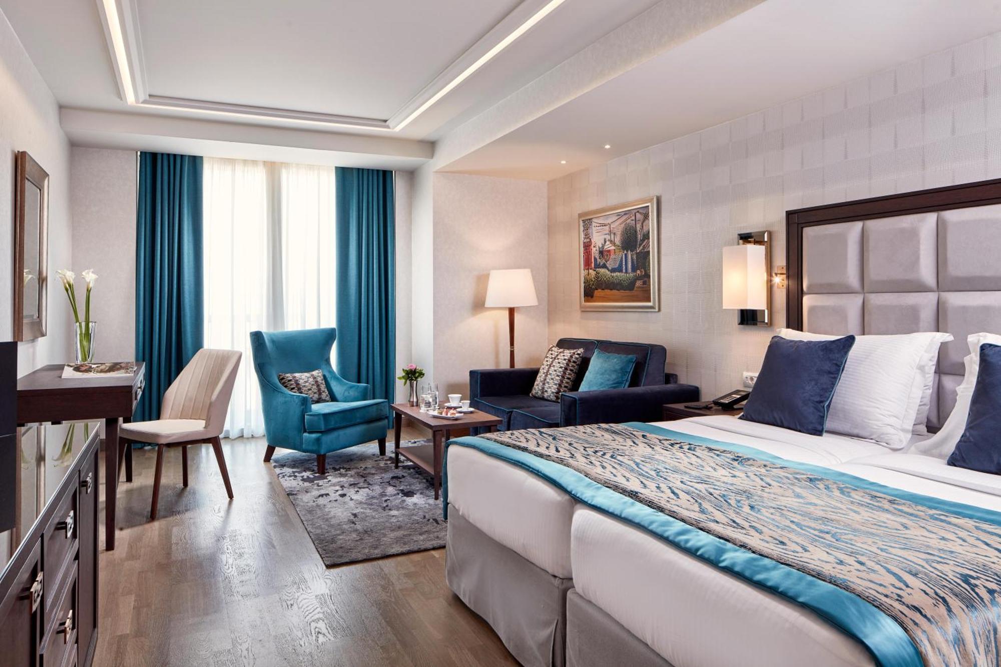 فندق إلكترا متروبوليس أثينا المظهر الخارجي الصورة The photo shows a modern hotel room featuring a double bed with a decorative blue throw at the foot. On one side, there are two large windows with turquoise curtains letting in natural light. There's a comfortable seating area with a blue chair, a sm