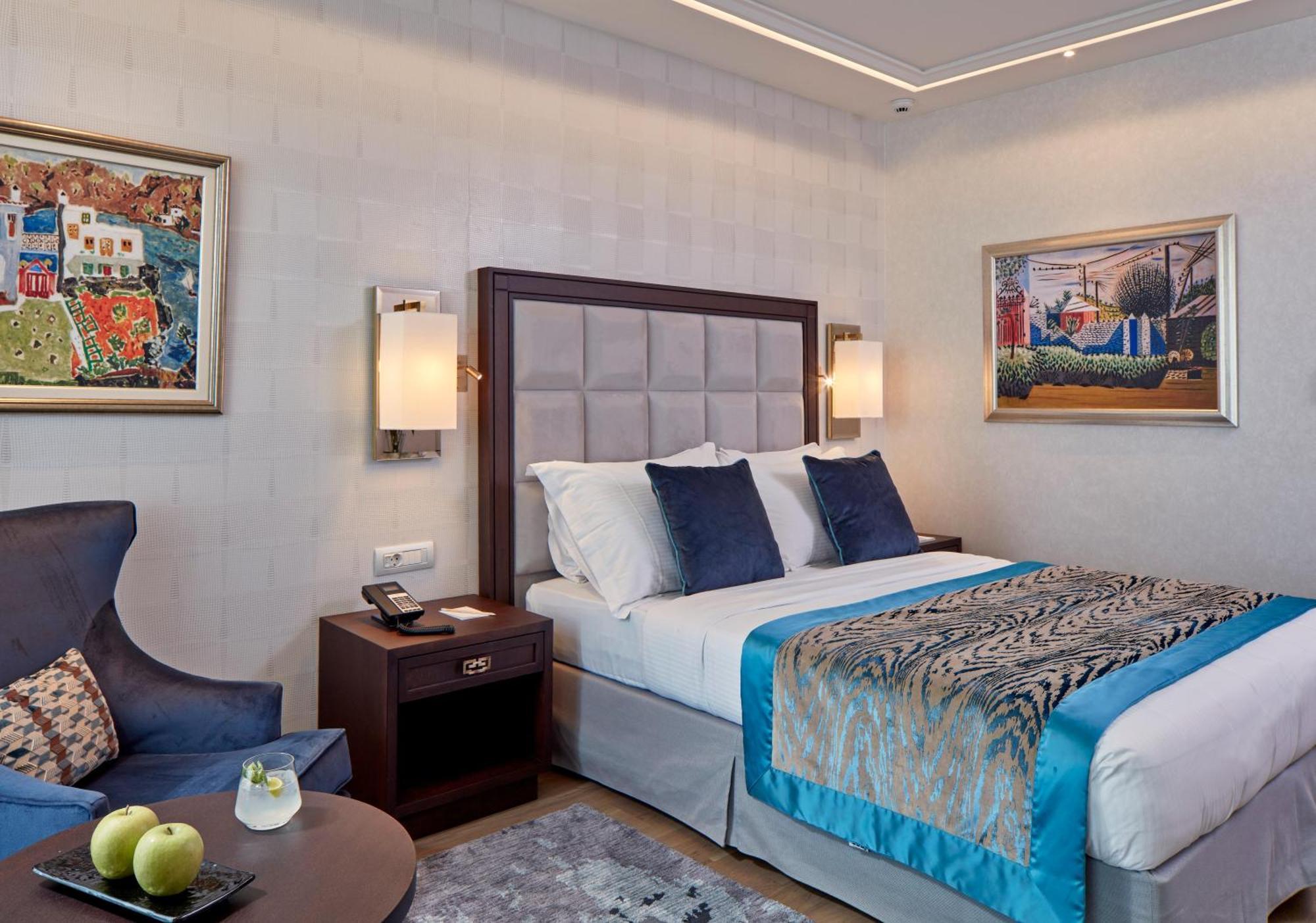 فندق إلكترا متروبوليس أثينا المظهر الخارجي الصورة The photo shows a modern hotel room featuring a double bed with decorative pillows. The bed has a stylish headboard, and the bedding includes a patterned throw. Beside the bed, there are bedside lamps and a wooden nightstand with a phone. 

On the le