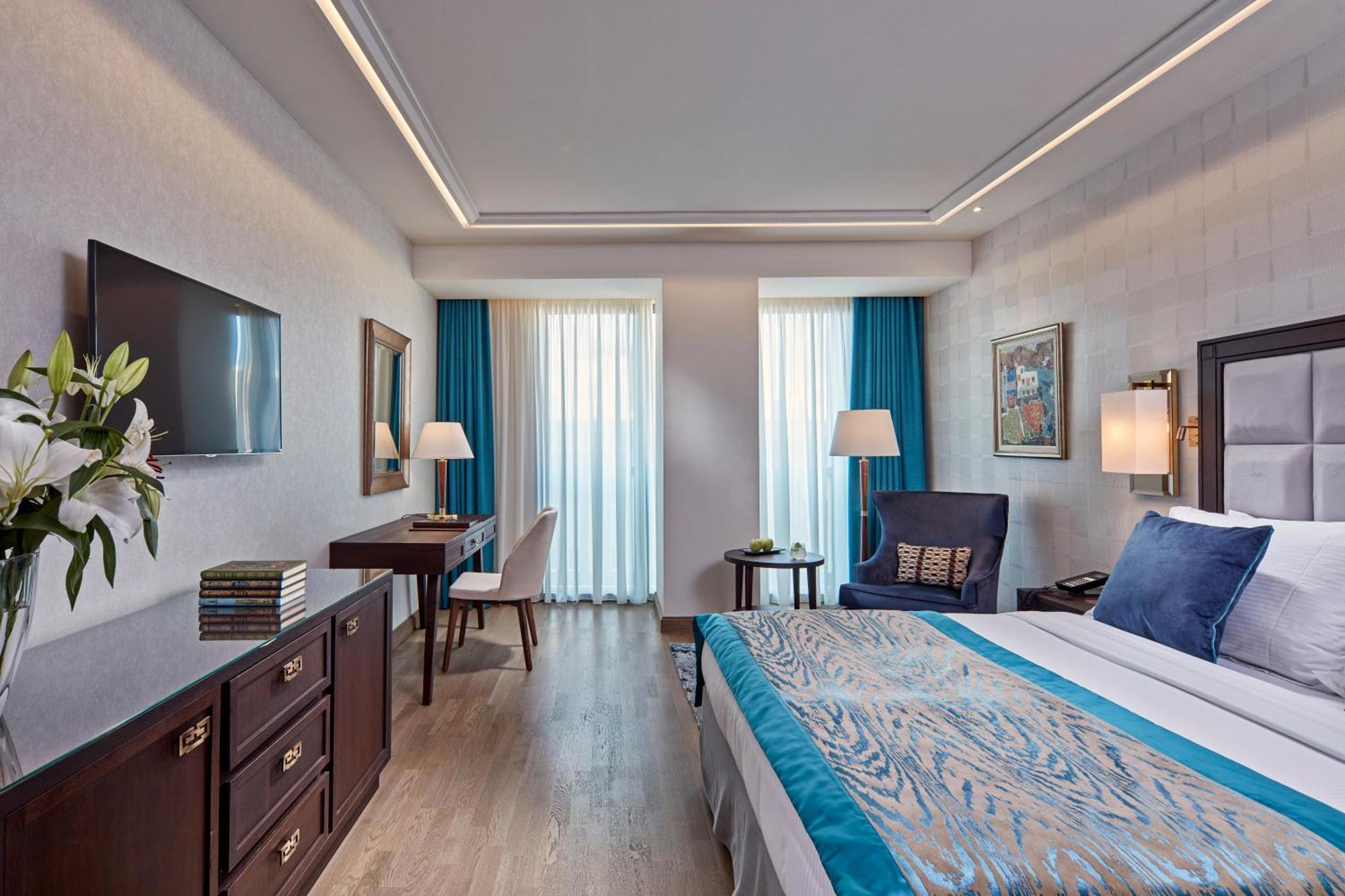 فندق إلكترا متروبوليس أثينا المظهر الخارجي الصورة The photo shows a well-decorated hotel room featuring a large bed with a blue and patterned bedspread. There are two windows with light blue curtains that allow natural light to fill the space. A flat-screen TV is mounted on the wall opposite the bed