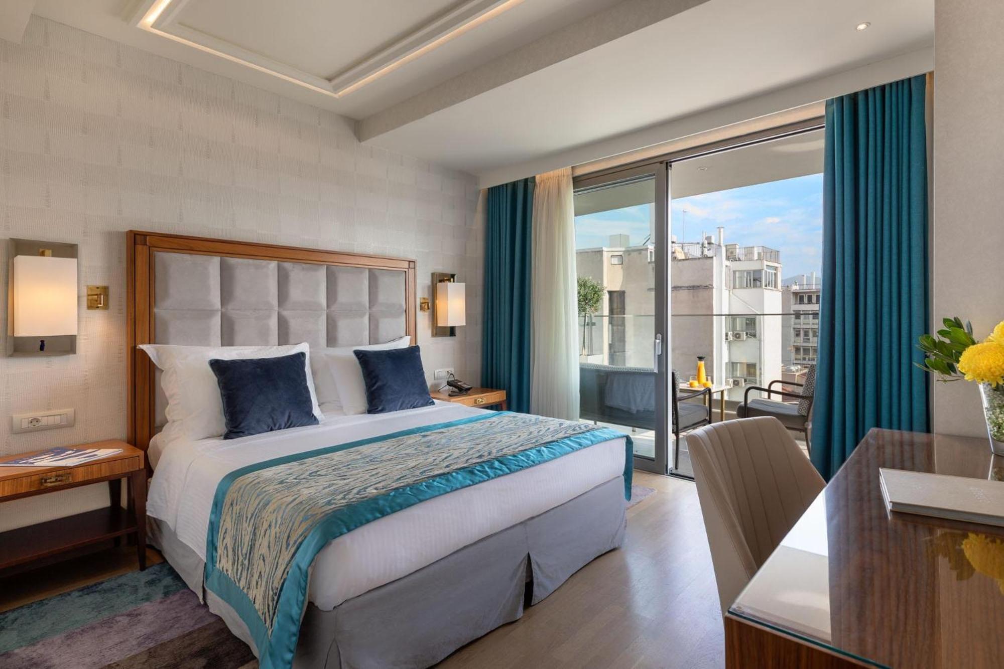 فندق إلكترا متروبوليس أثينا المظهر الخارجي الصورة The photo shows a modern hotel room featuring a queen-sized bed with a light-colored headboard. The bed is adorned with decorative pillows and a stylish throw blanket. There are two bedside lamps on either side of the bed. A set of floor-to-ceiling w