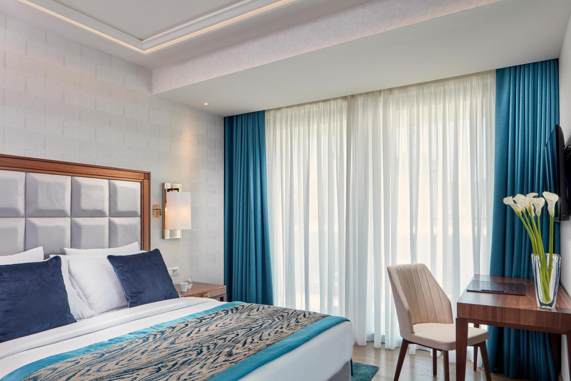 فندق إلكترا متروبوليس أثينا المظهر الخارجي الصورة The photo shows a modern hotel room featuring a large bed with decorative pillows and a stylish headboard. The bedding is accented with a patterned throw blanket. There are floor-to-ceiling windows with sheer and solid blue curtains allowing natural 