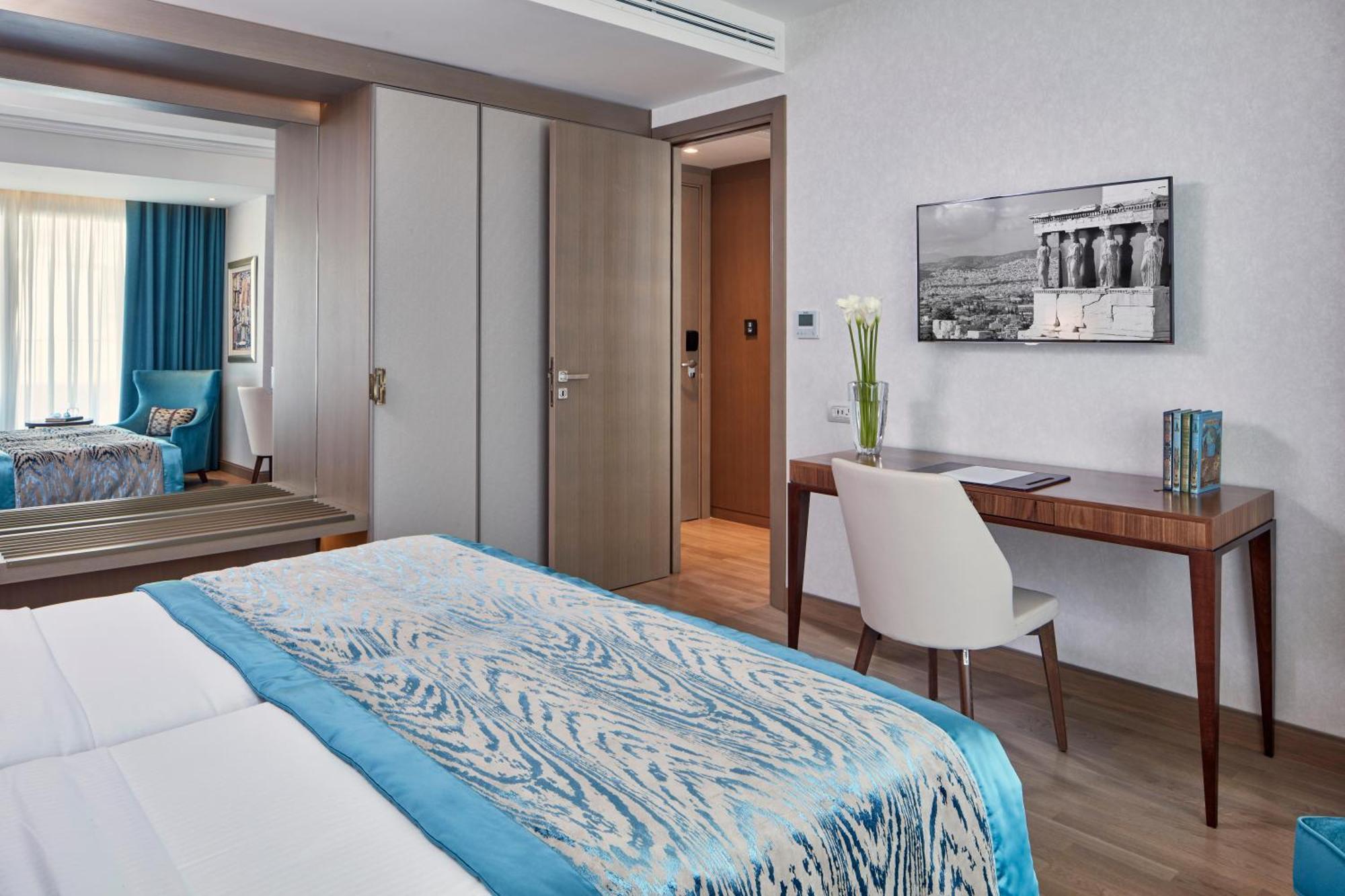 فندق إلكترا متروبوليس أثينا المظهر الخارجي الصورة The photo shows a modern hotel room with two beds covered in light-colored linens that have a blue patterned throw or blanket. There's a wooden desk and a white chair situated next to the wall, with a vase of flowers on the desk and a framed black-an