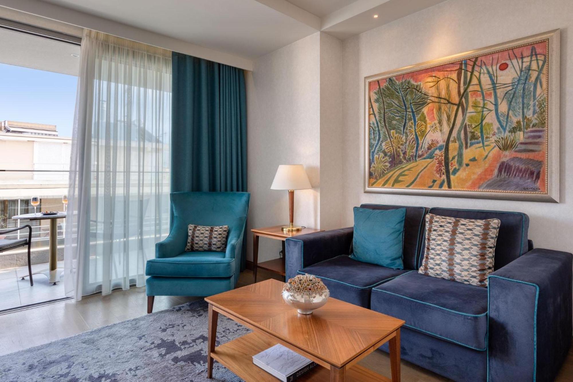 فندق إلكترا متروبوليس أثينا المظهر الخارجي الصورة The photo shows a modern living room area. There is a blue sofa with decorative pillows and a wooden coffee table in front of it. A light blue armchair is positioned next to the sofa. A painting with vibrant colors depicting a natural scene is hangin