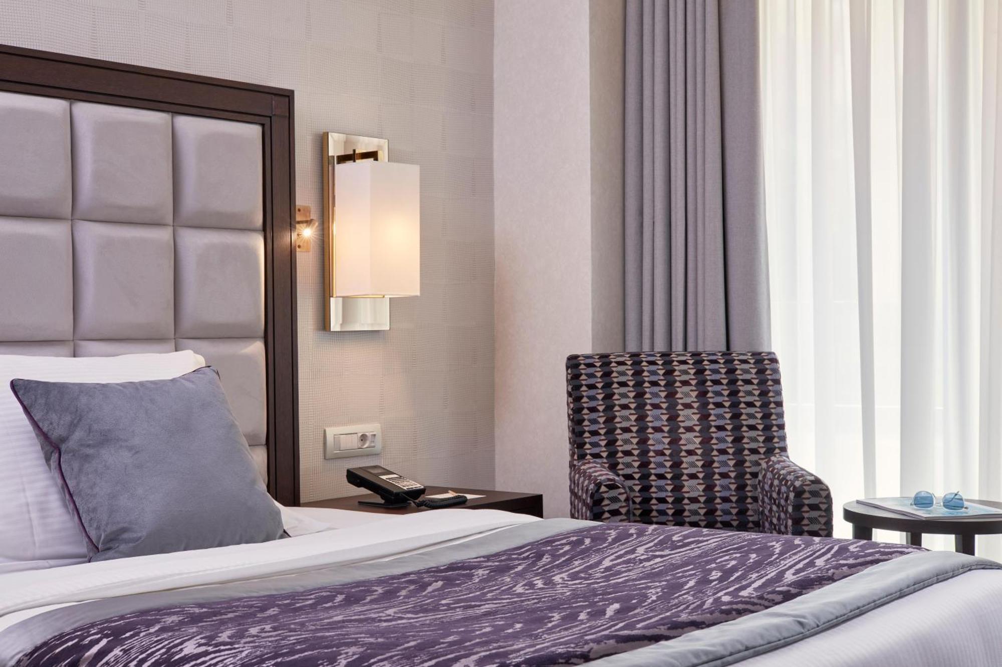 فندق إلكترا متروبوليس أثينا المظهر الخارجي الصورة The photo shows a modern hotel room featuring a neatly made bed with a dark purple throw blanket and large pillows. Behind the bed, there is a stylish headboard with a tufted design. A soft, ambient light fixture is mounted on the wall beside the bed