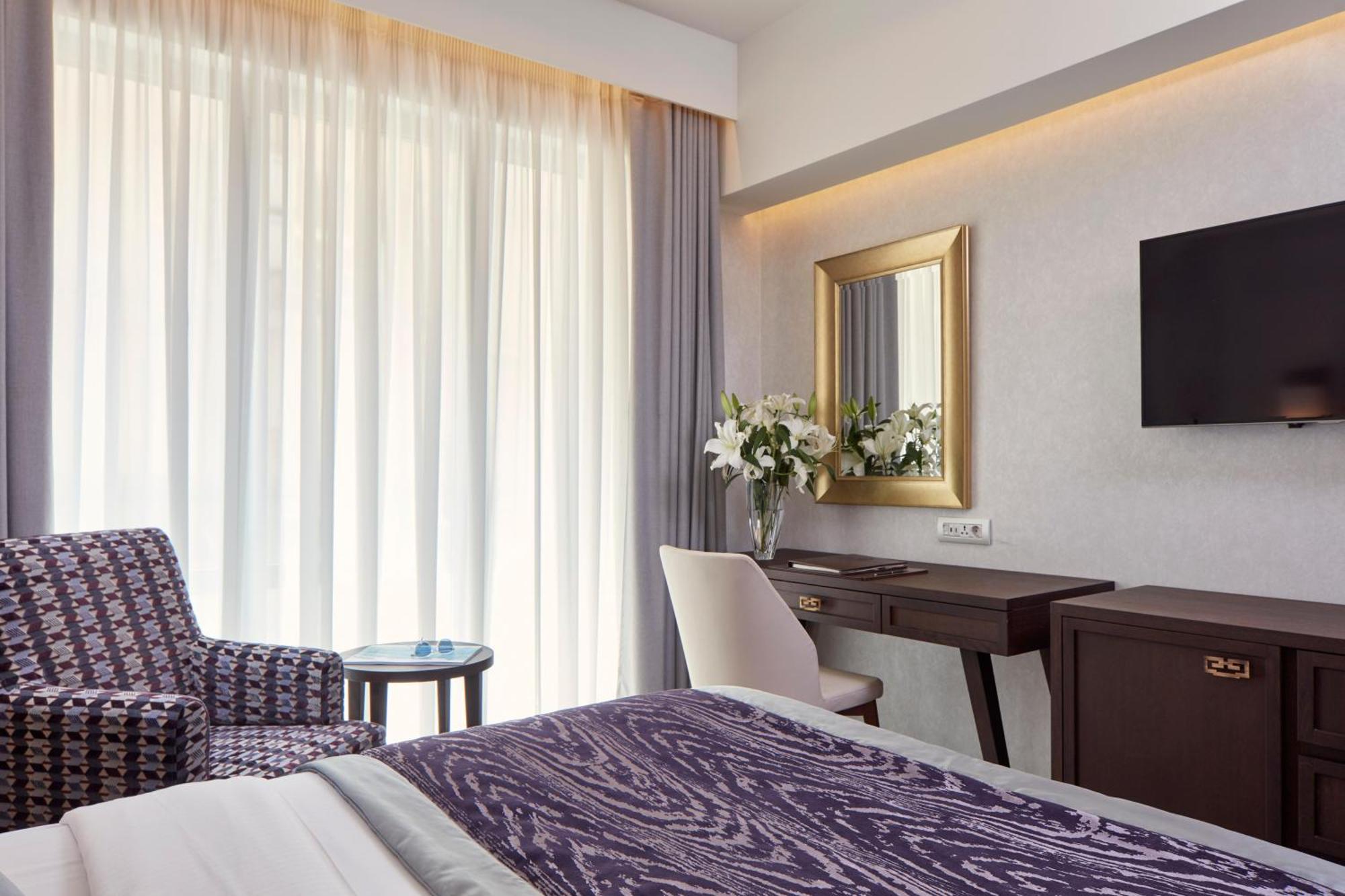 فندق إلكترا متروبوليس أثينا المظهر الخارجي الصورة The photo shows a well-furnished hotel room. On the left, there is a patterned chair with a small round table next to it. The room features large windows covered with sheer curtains that allow natural light to fill the space. In the center, there is 