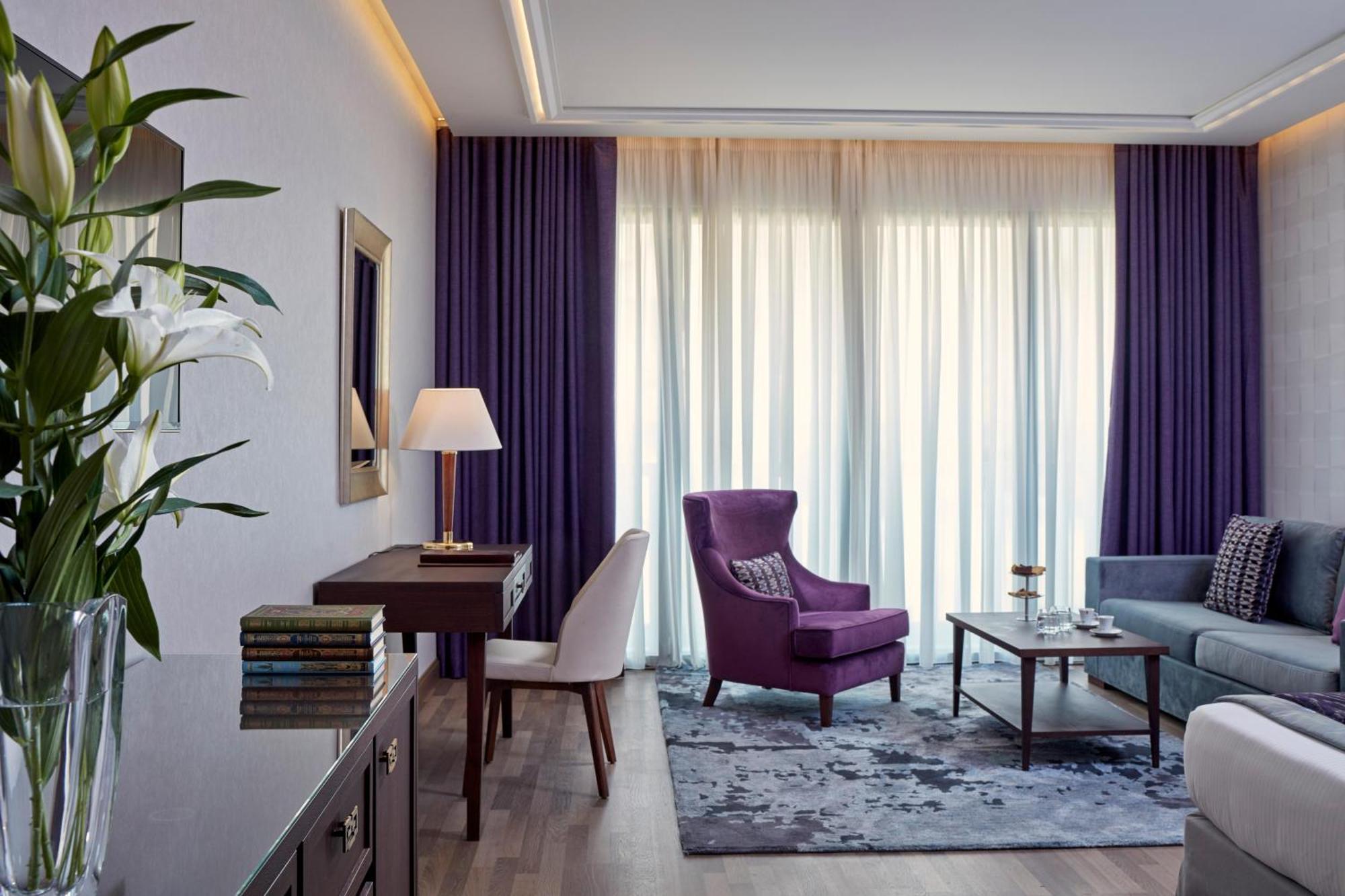 فندق إلكترا متروبوليس أثينا المظهر الخارجي الصورة The photo depicts a stylish and modern living space. On one side, there's a plush purple armchair and a small wooden side table with a lamp. Nearby, a light-colored sofa is placed with a coffee table in front of it. The room features large windows co