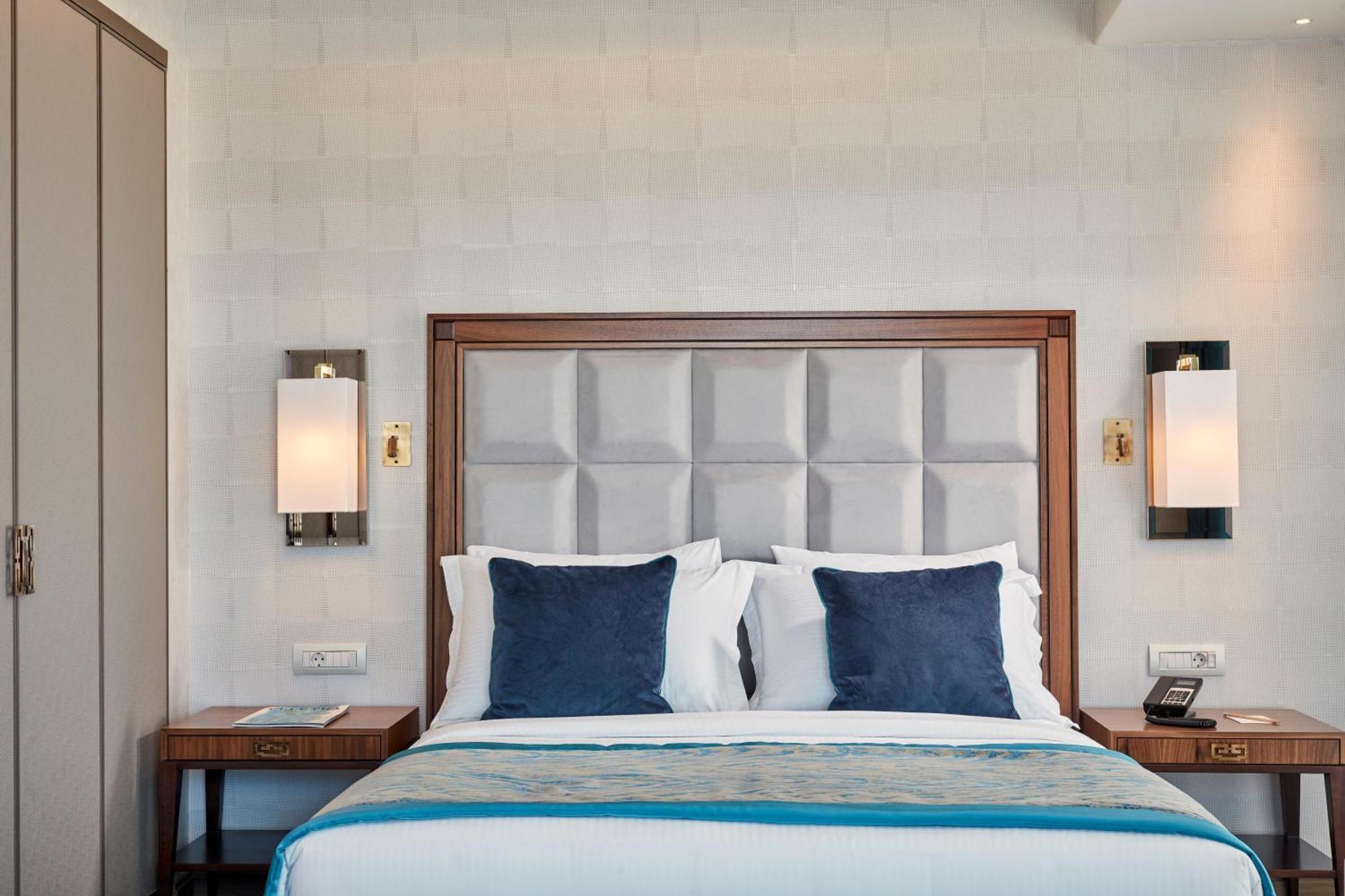 فندق إلكترا متروبوليس أثينا المظهر الخارجي الصورة The photo shows a well-designed hotel room with a cozy bed as the focal point. The bed has a soft, padded headboard in a light color, paired with four decorative pillows—two in a dark blue shade and two in white. On either side of the bed, there are 