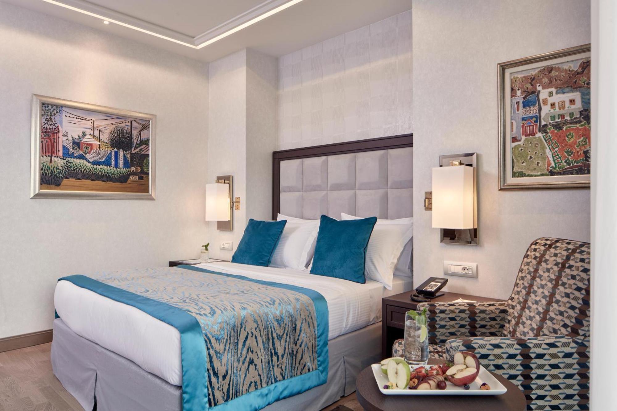 فندق إلكترا متروبوليس أثينا المظهر الخارجي الصورة The photo shows a hotel room featuring a queen-sized bed with a light blue and patterned bedspread. The bed has white pillows with two accent pillows in turquoise. Beside the bed, there are contemporary lamps mounted on the wall. 

To the side, there