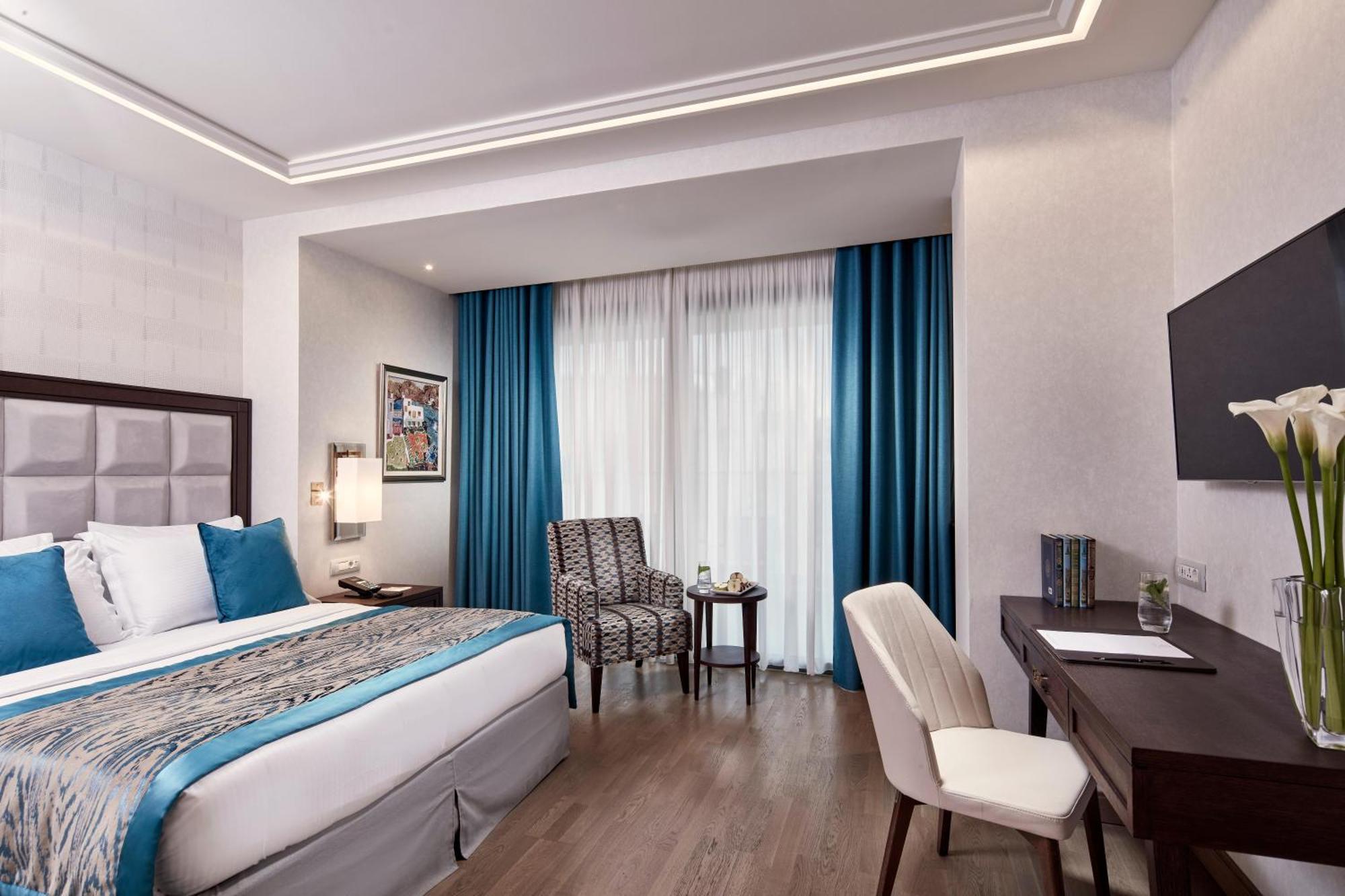 فندق إلكترا متروبوليس أثينا المظهر الخارجي الصورة The photo shows a well-decorated hotel room. It features a large bed with a blue and neutral colored bedding set. There are two blue curtains that frame a window, allowing natural light to enter the space. Next to the bed, there's a small chair and t