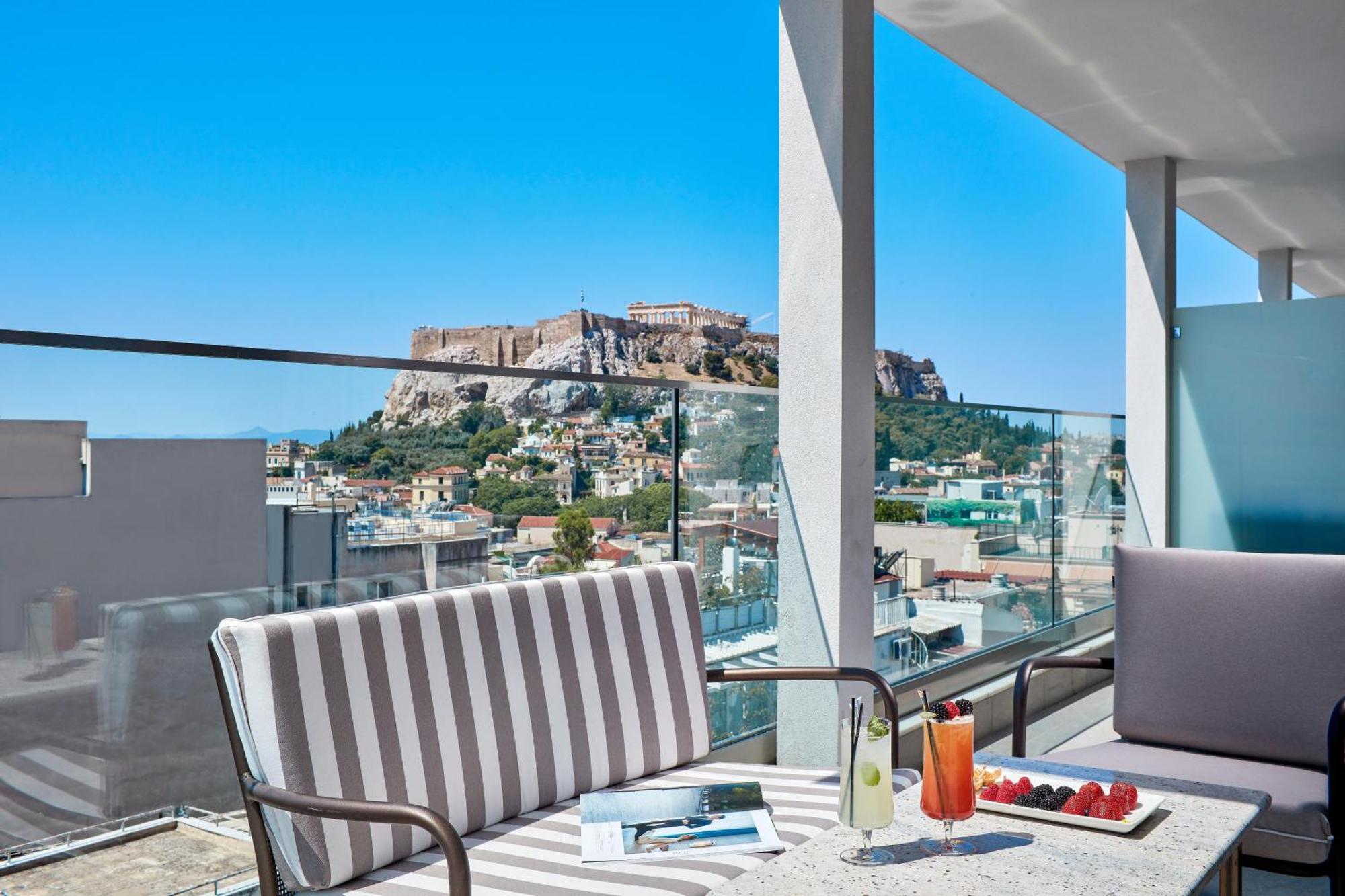 فندق إلكترا متروبوليس أثينا المظهر الخارجي الصورة The photo features a modern terrace or balcony with stylish seating arrangements. There are two cushioned chairs, one with striped fabric. A table in front holds a beverage, possibly a cocktail, and some fresh strawberries. The backdrop showcases a b