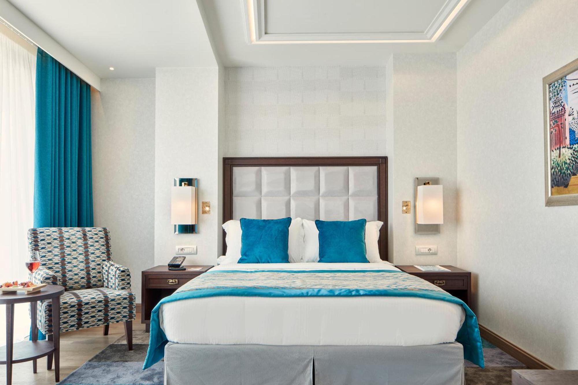 فندق إلكترا متروبوليس أثينا المظهر الخارجي الصورة The photo shows a modern hotel room. In the center, there is a large bed with a white and light blue bedding set. The headboard is upholstered in a light gray material. On either side of the bed, there are wall-mounted lamps. There is a stylish chair