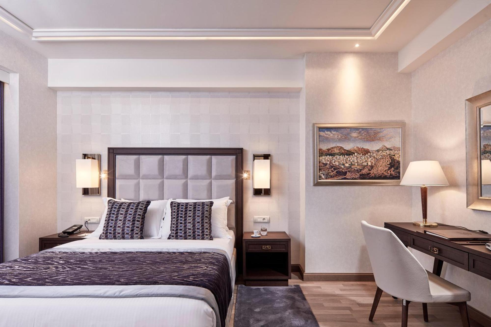 فندق إلكترا متروبوليس أثينا المظهر الخارجي الصورة The photo shows a stylish hotel room featuring a double bed with a plush headboard and decorative pillows. On either side of the bed, there are wall-mounted lamps providing soft lighting. There's a small nightstand beside the bed and a modern desk ch