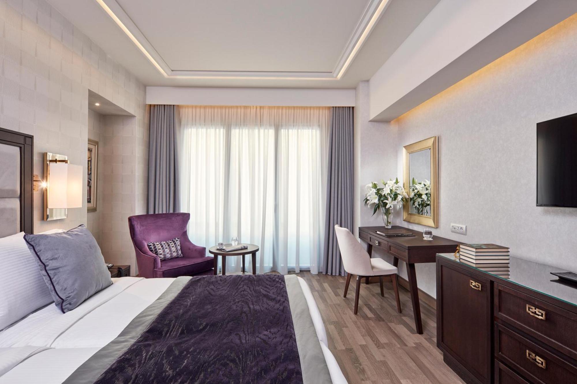 فندق إلكترا متروبوليس أثينا المظهر الخارجي الصورة The photo shows a well-decorated hotel room with a modern and elegant design. The room features a large bed with a dark purple throw blanket and plush pillows. There is a cozy purple armchair beside a small round table. A stylish white chair is posit