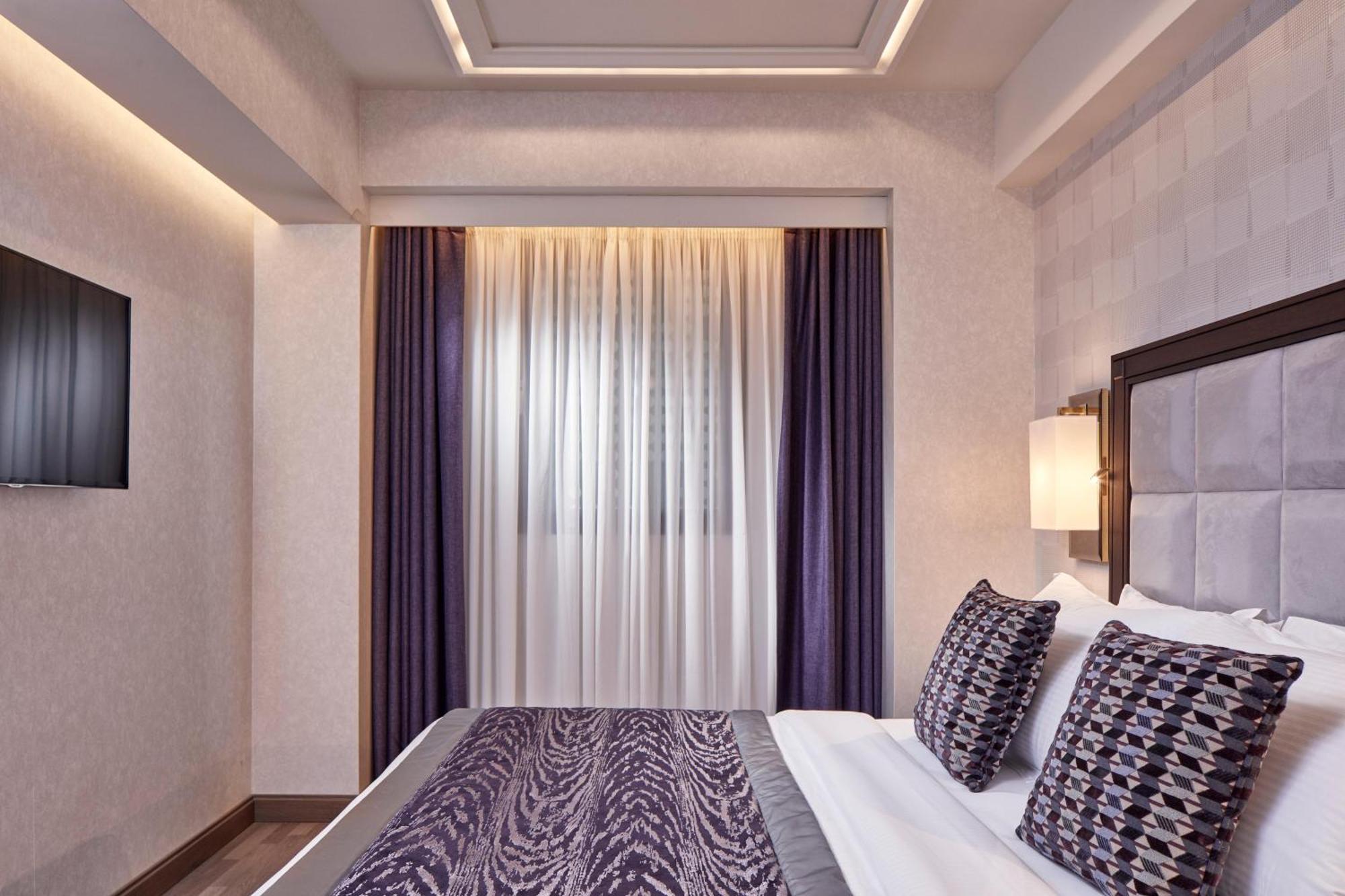 فندق إلكترا متروبوليس أثينا المظهر الخارجي الصورة The photo shows a modern bedroom featuring a bed with patterned pillows and a decorative comforter. The room has soft lighting, with a wall-mounted TV visible. There are large curtains that allow natural light to filter in, and the walls have a subtl