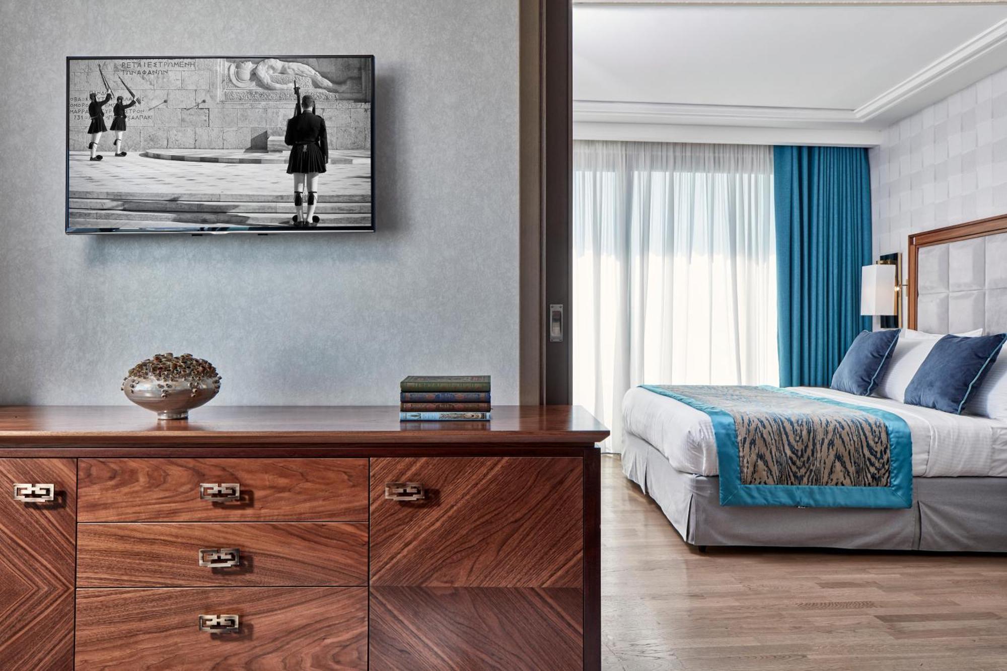 فندق إلكترا متروبوليس أثينا المظهر الخارجي الصورة The photo shows a modern, stylish hotel room or suite. In the foreground, there is a wooden dresser with several drawers, and on top, there is a decorative piece, a few books, and a television mounted on the wall displaying a black-and-white image of