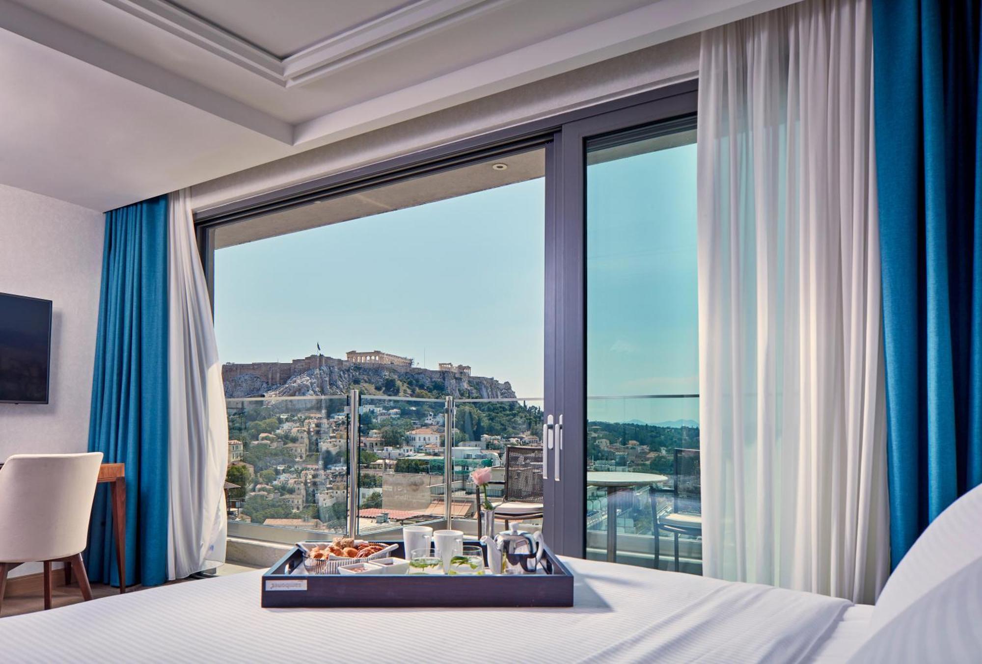 فندق إلكترا متروبوليس أثينا المظهر الخارجي الصورة The photo shows a modern hotel room with a large window offering a view of a historical site, likely a fortress or ancient ruins, situated on a hill. The interior features a neatly made bed with a tray placed on it, displaying various items that coul