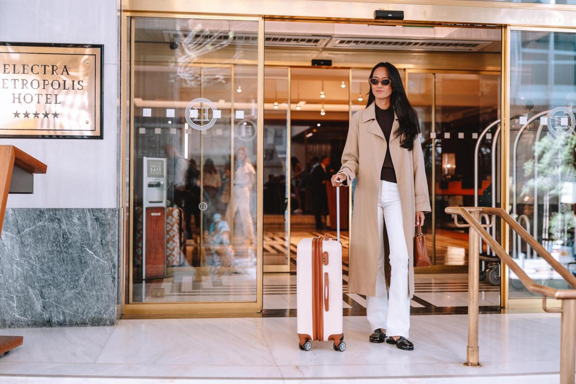 فندق إلكترا متروبوليس أثينا المظهر الخارجي الصورة The photo shows a fashionable individual standing outside a hotel entrance. The person is wearing a long beige coat over a dark top, paired with white pants. They are holding a stylish suitcase with a wooden design. The hotel has large glass doors, a