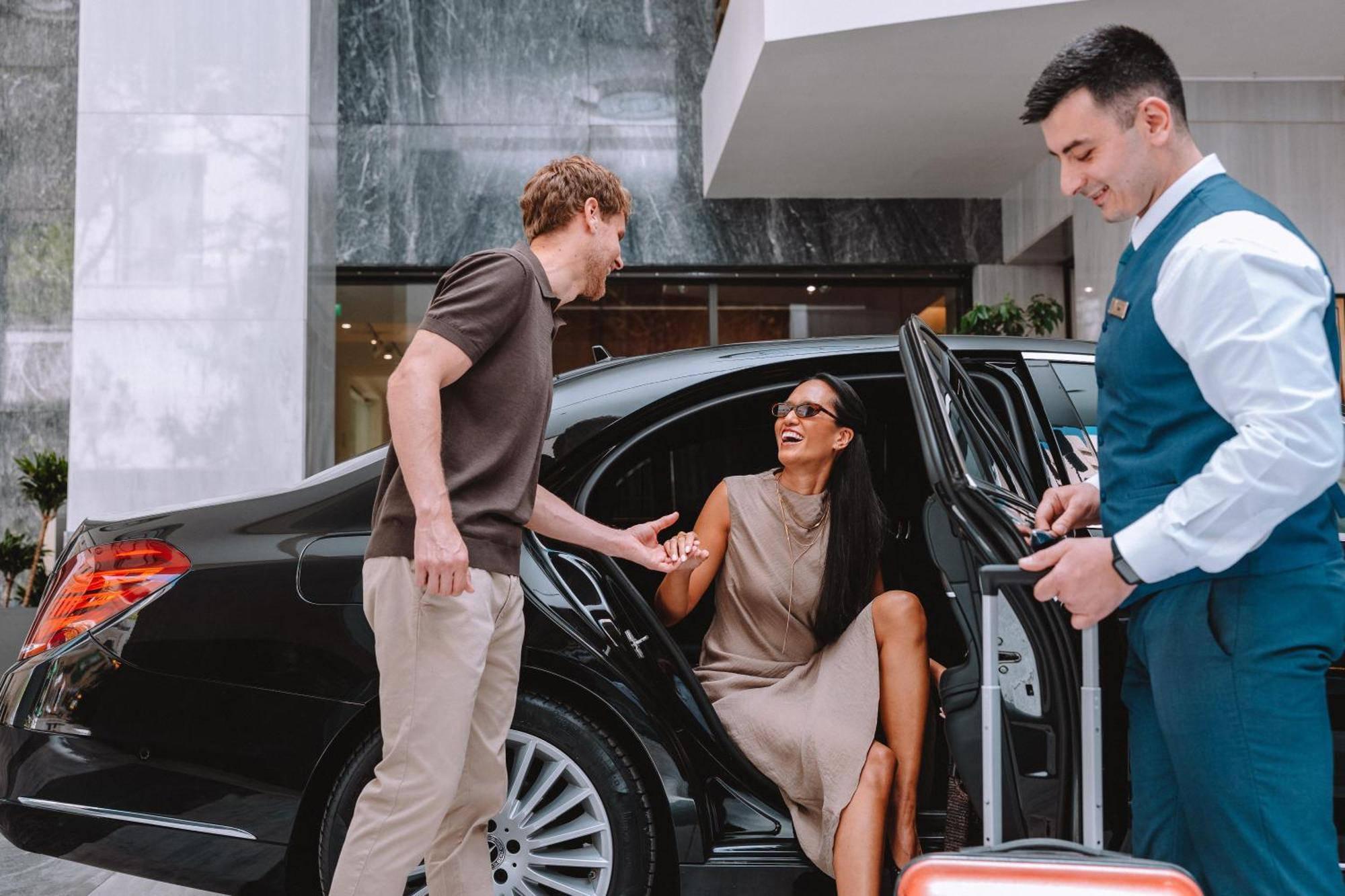 فندق إلكترا متروبوليس أثينا المظهر الخارجي الصورة The photo shows a scene outside a luxury hotel. A man is standing next to an open car door, interacting with a woman who is seated inside the car. The woman, wearing sunglasses and a stylish outfit, appears to be smiling and enjoying the moment. A ho