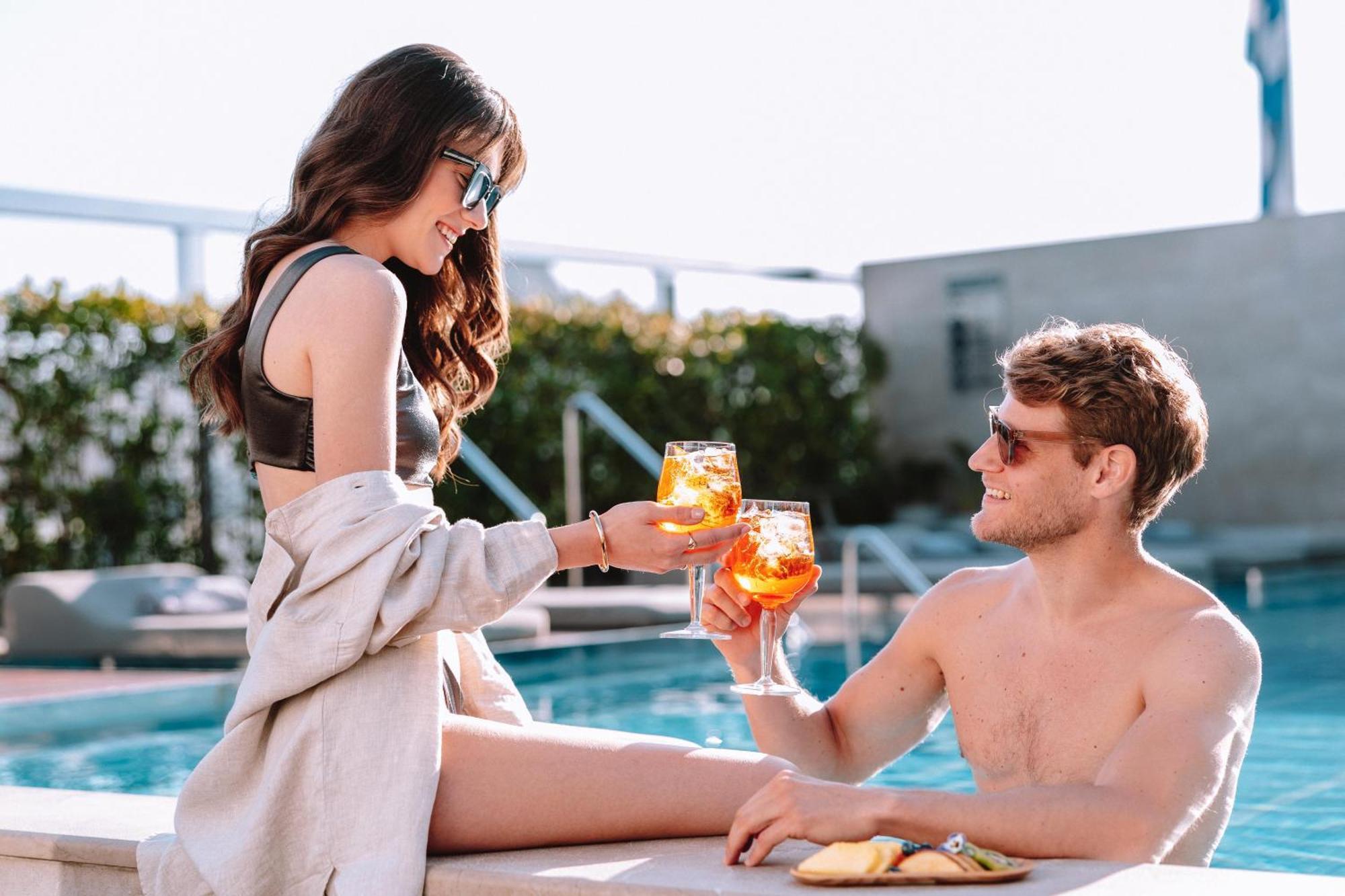 فندق إلكترا متروبوليس أثينا المظهر الخارجي الصورة The photo shows a pair enjoying a vibrant moment by a pool. A woman, sitting on the edge, is wearing stylish sunglasses and a bikini, and she is handing a drink to a man sitting beside her. He is shirtless, also wearing sunglasses, and is smiling whi