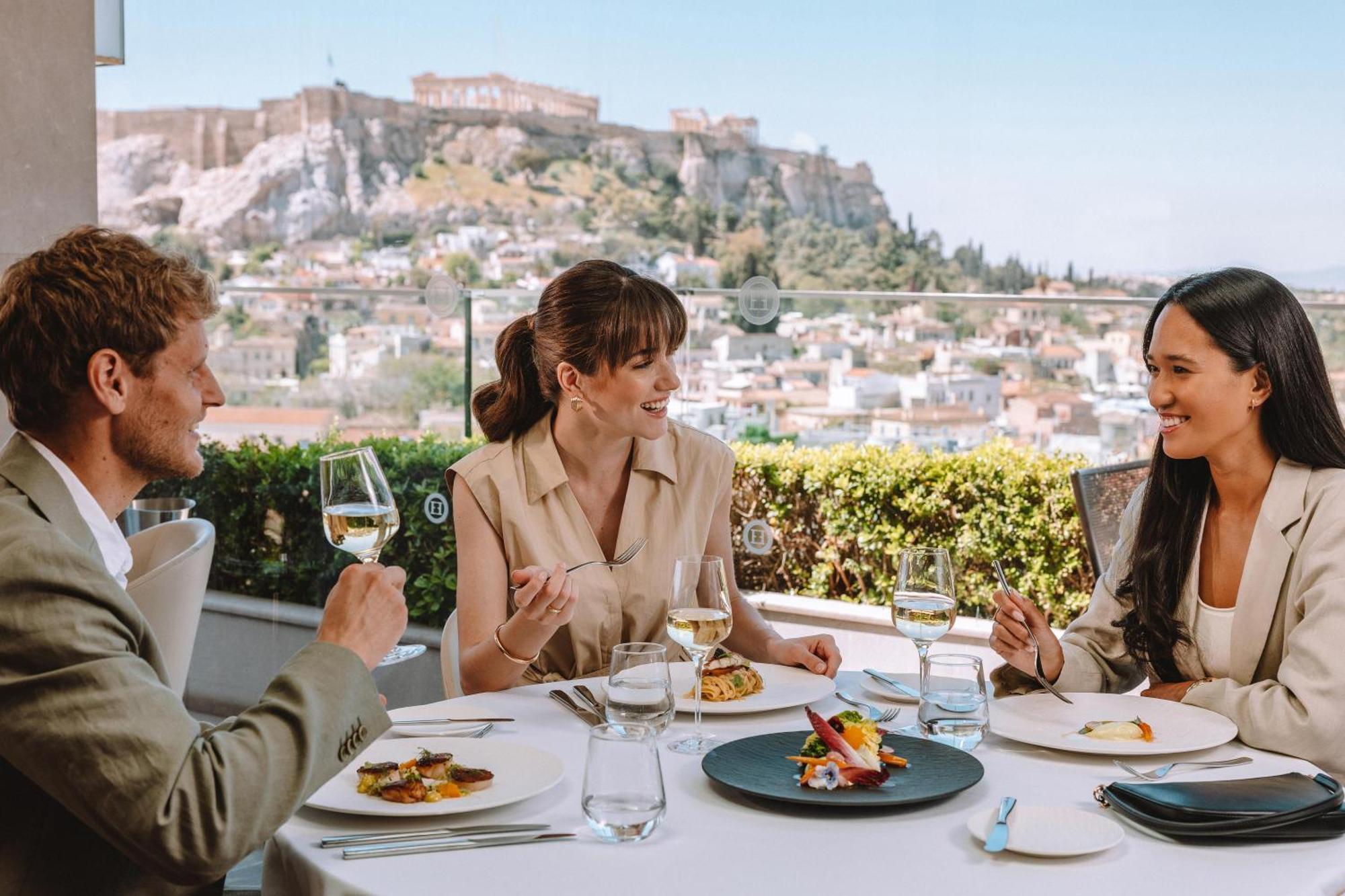 فندق إلكترا متروبوليس أثينا المظهر الخارجي الصورة The photo depicts three people enjoying a meal at an outdoor restaurant with a scenic view in the background. There are two women and one man seated at a table that includes several plates of food and glasses of wine. The atmosphere appears relaxed a